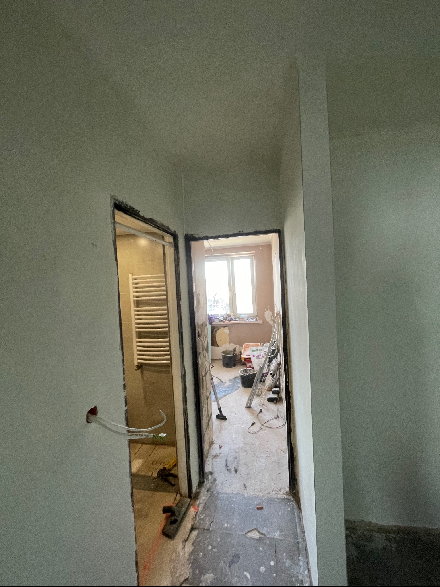 Wnętrze w trakcie remontu: widok na dwa pomieszczenia przez surowe otwory drzwiowe. Ściany po szpachlowaniu, widoczne przewody elektryczne i narzędzia. Podłoga częściowo wykończona.