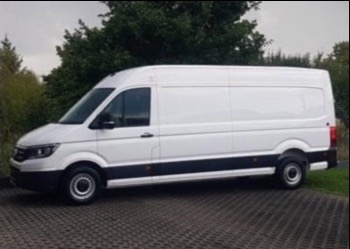 Biały, długi bus dostawczy marki MAN na brukowanym podjeździe, z zielenią w tle. Widok z boku, lekko od dołu, podkreślający jego rozmiar.