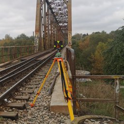 Geo-SKan Usługi Geodezyjno-Kartograficzne Stanisław Kurdybacha - Żółty niwelator na trójnogu ustawiony na betonowym fundamencie, na tle zardzewiałego mostu kolejowego z graffiti i jesiennej zieleni. Pomiar geodezyjny na moście.