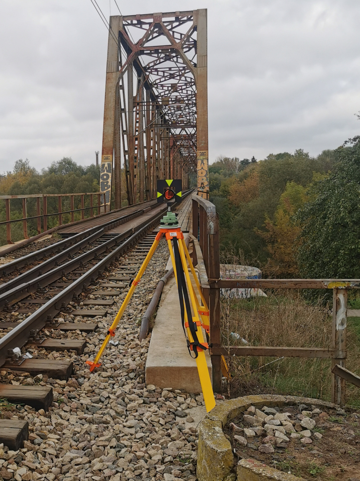 Żółty niwelator na trójnogu ustawiony na betonowym fundamencie, na tle zardzewiałego mostu kolejowego z graffiti i jesiennej zieleni. Pomiar geodezyjny na moście.