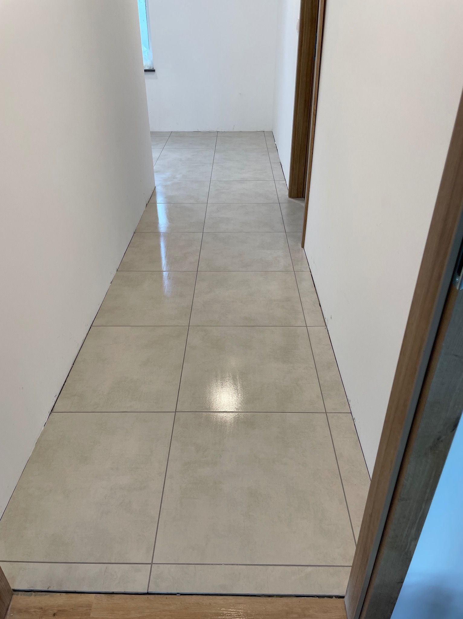 Położone, jasne płytki podłogowe w korytarzu, widoczne z perspektywy podłogi. Świeżo wykończone, błyszczące. Fragment ościeżnicy po prawej stronie kadru.