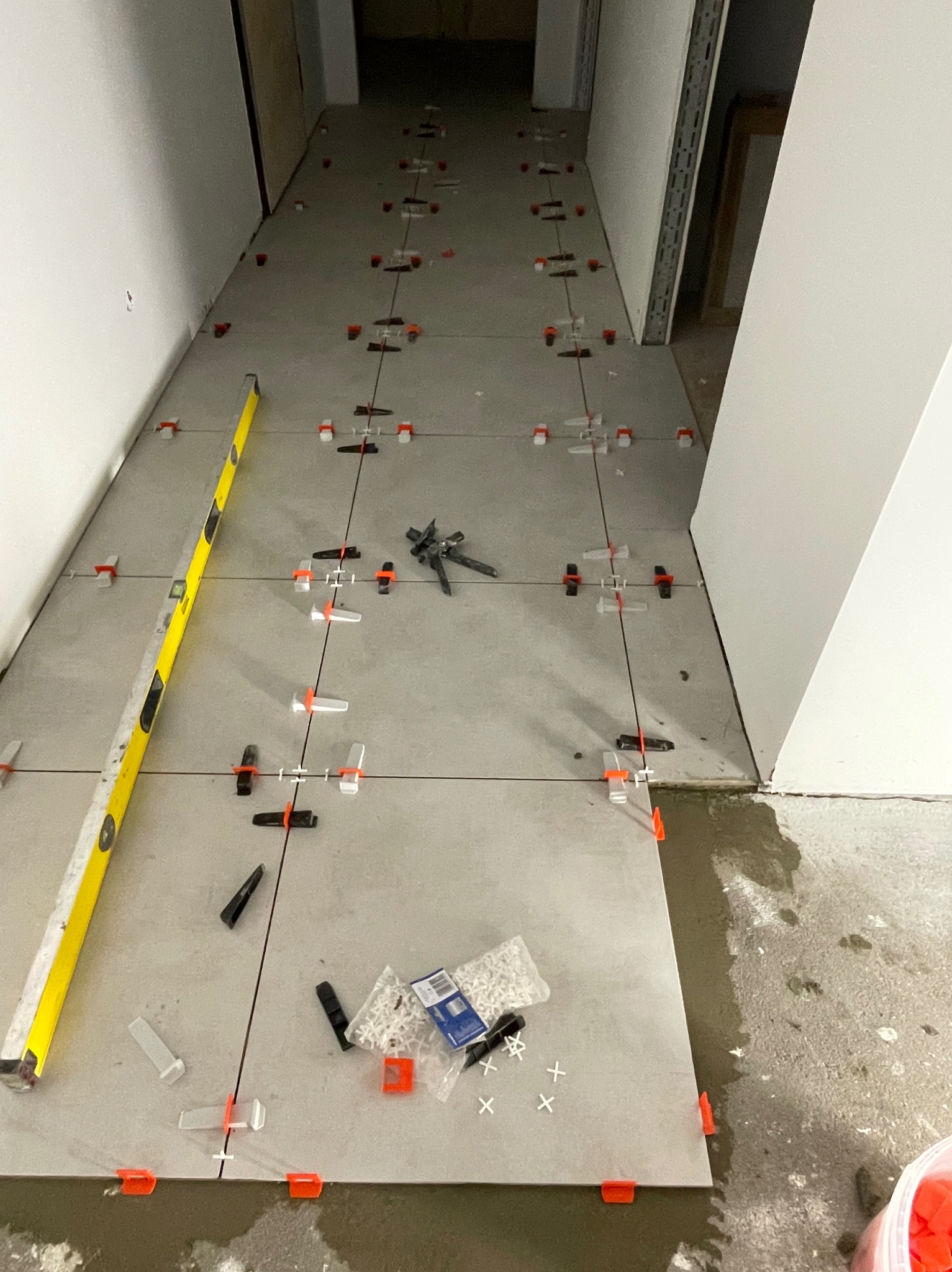Układanie szarych płytek podłogowych w korytarzu z użyciem poziomicy, klipsów i krzyżyków dystansowych. Widoczne narzędzia glazurnicze i opakowanie z krzyżykami.