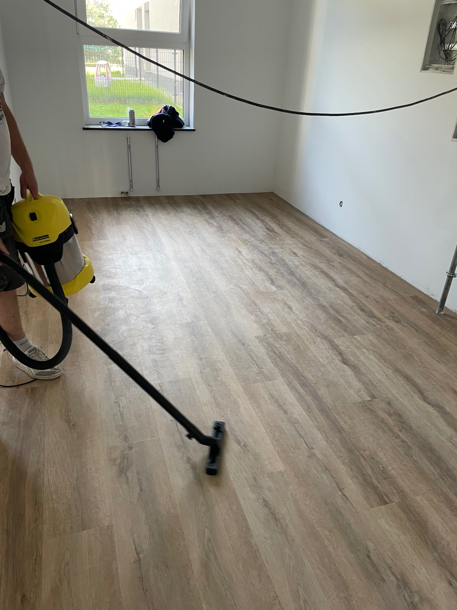 Jasne panele podłogowe po montażu, czyszczone odkurzaczem przemysłowym. Widoczne okno i fragment instalacji elektrycznej na ścianie. Ujęcie z bliska, z boku.