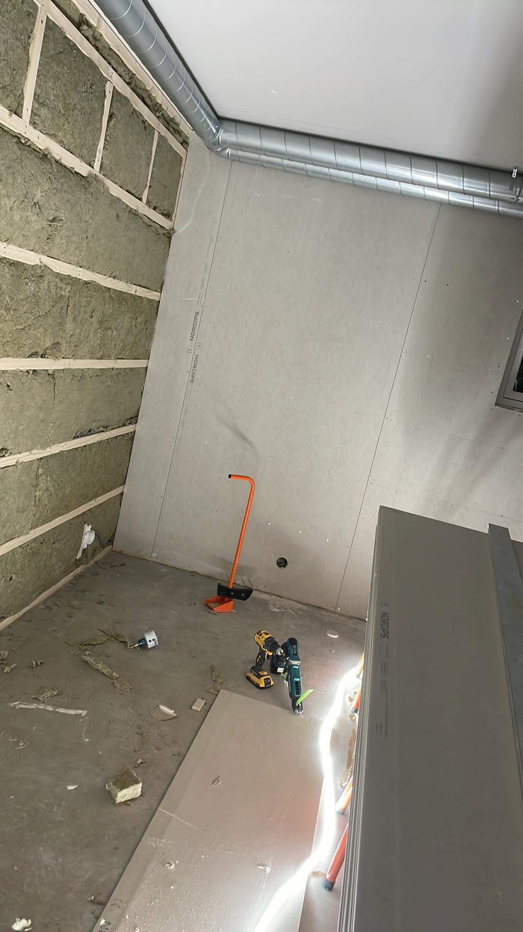 Ocieplanie ścian wełną mineralną i montaż płyt gipsowo-kartonowych w pomieszczeniu z wentylacją. Widoczne narzędzia i resztki materiałów na podłodze.