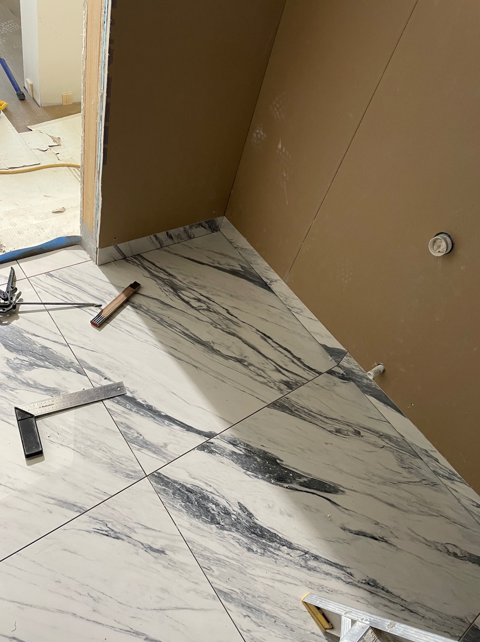 Ułożone marmurowe płytki podłogowe w łazience, widoczne narzędzia glazurnicze: kątownik, miarka składana. Ściany przygotowane pod wykończenie, widoczne instalacje.