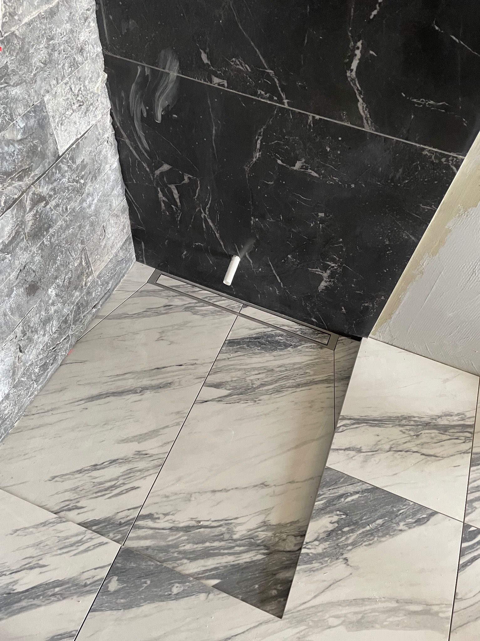 Ukończona łazienka z marmurowymi płytkami na podłodze i ścianach, czarny marmur kontrastuje z szarym kamieniem. Widoczny odpływ liniowy.