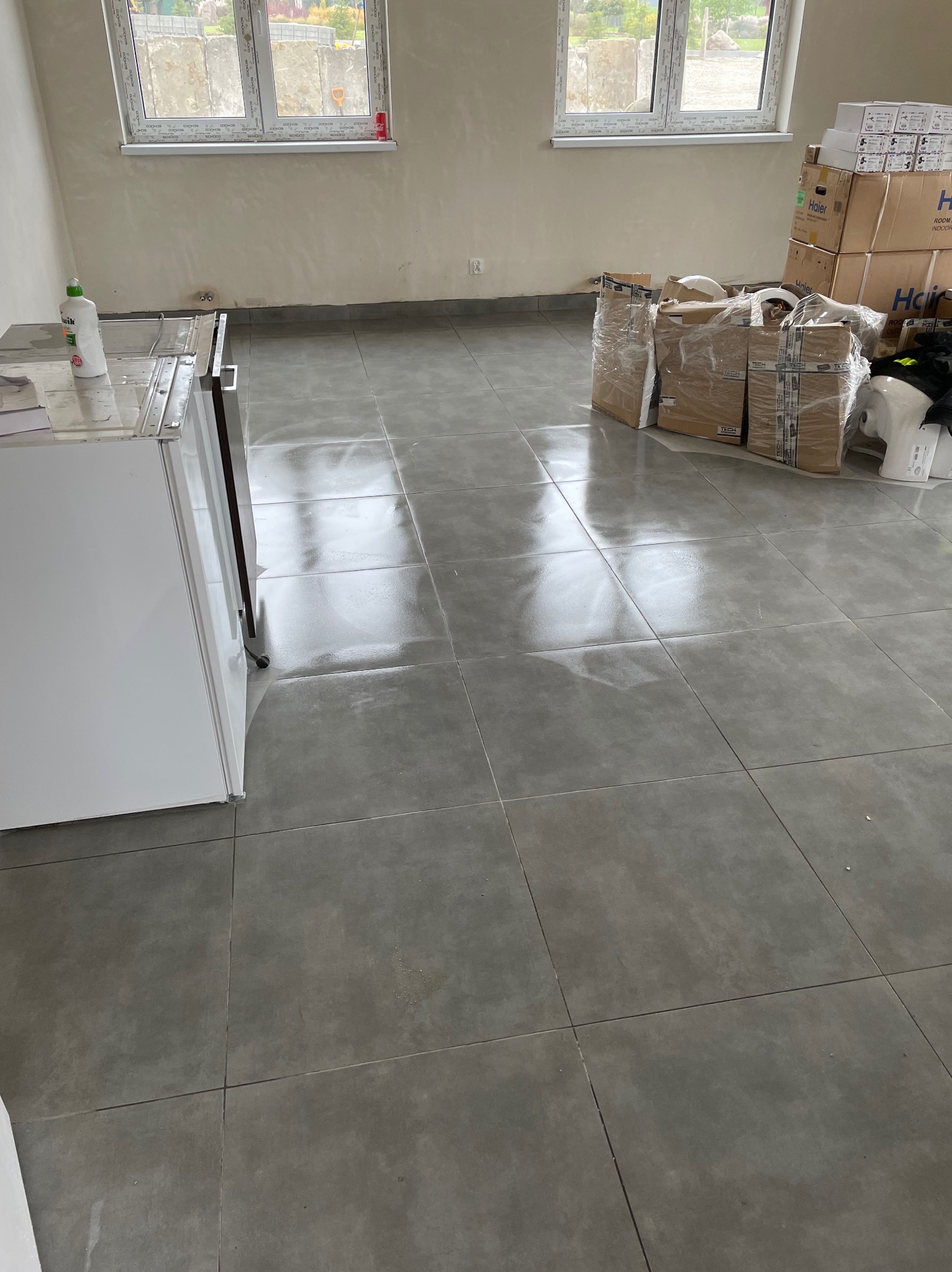 Ułożone, szare płytki podłogowe w nowym pomieszczeniu z oknami i stertą zapakowanych urządzeń. Widoczny fragment białej szafki i butelka płynu.