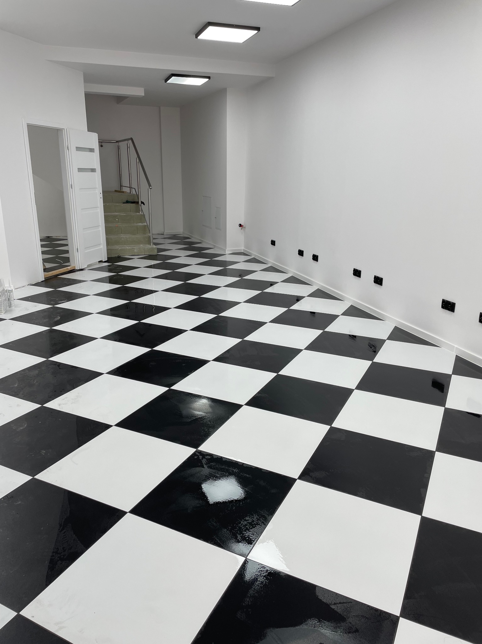 Błyszcząca, czarno-biała podłoga w szachownicę w pustym, białym pomieszczeniu z widocznymi schodami i gniazdkami elektrycznymi wzdłuż ściany.