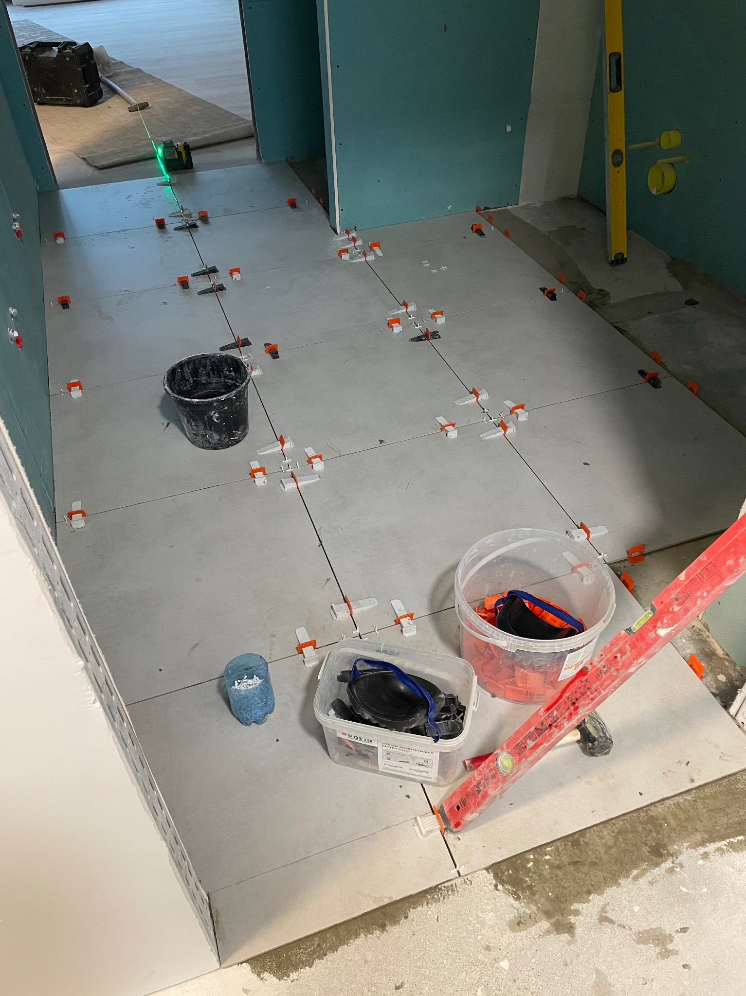 Układanie jasnych płytek podłogowych z użyciem poziomicy laserowej i klipsów poziomujących. Widoczne narzędzia glazurnicze i fragment ściany z płyt karton-gips.
