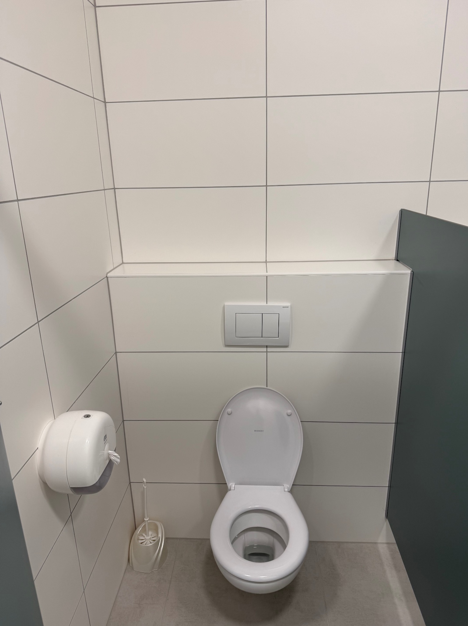 Biała toaleta z otwartą klapą w nowoczesnej łazience wyłożonej białymi płytkami. Widoczny dozownik papieru i szczotka do WC. Minimalistyczny design.