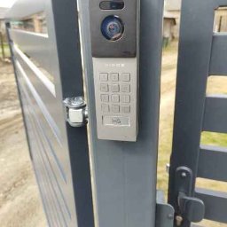 ELSMART Krzysztof Różyło - Szary wideofon Vidos z klawiaturą numeryczną zamontowany na słupku ogrodzenia panelowego. Widoczny fragment furtki i otoczenie domu.