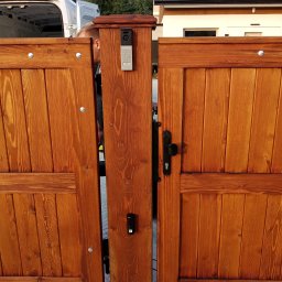 ELSMART Krzysztof Różyło - Drewniana brama wjazdowa z domofonem i panelem numerycznym, widoczny fragment budynku w tle. Brama w kolorze ciepłego brązu, z widoczną strukturą drewna.