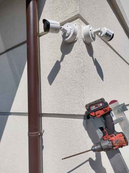 Montaż dwóch białych kamer monitoringu na elewacji budynku, widoczna wiertarka i drabina, brązowa rynna w tle. Ujęcie z dołu.