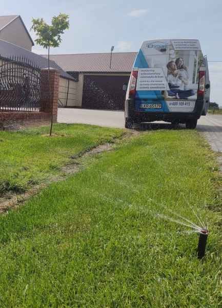 Działający system nawadniania trawnika z dyszą rozpylającą wodę. W tle budynek i samochód dostawczy z reklamą firmy.