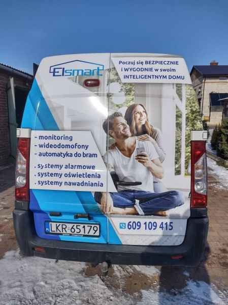 Tył furgonetki z reklamą firmy Elsmart: monitoring, wideodomofony, automatyka do bram, systemy alarmowe, oświetlenia i nawadniania. Numer telefonu i hasło: Inteligentny dom.