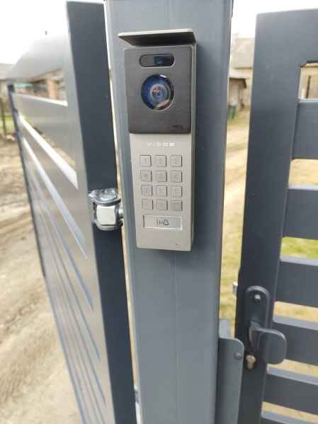 Szary wideofon Vidos z klawiaturą numeryczną zamontowany na słupku ogrodzenia panelowego. Widoczny fragment furtki i otoczenie domu.