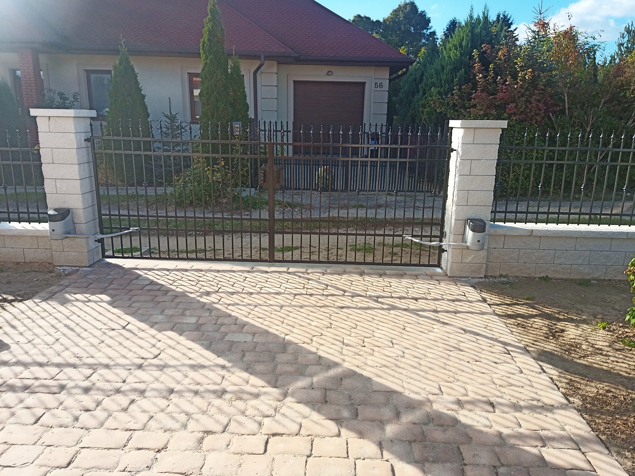 Automatyczna brama przesuwna z metalowymi sztachetami i kamiennymi słupkami, widok z zewnątrz na posesję z kostką brukową i domem w tle.