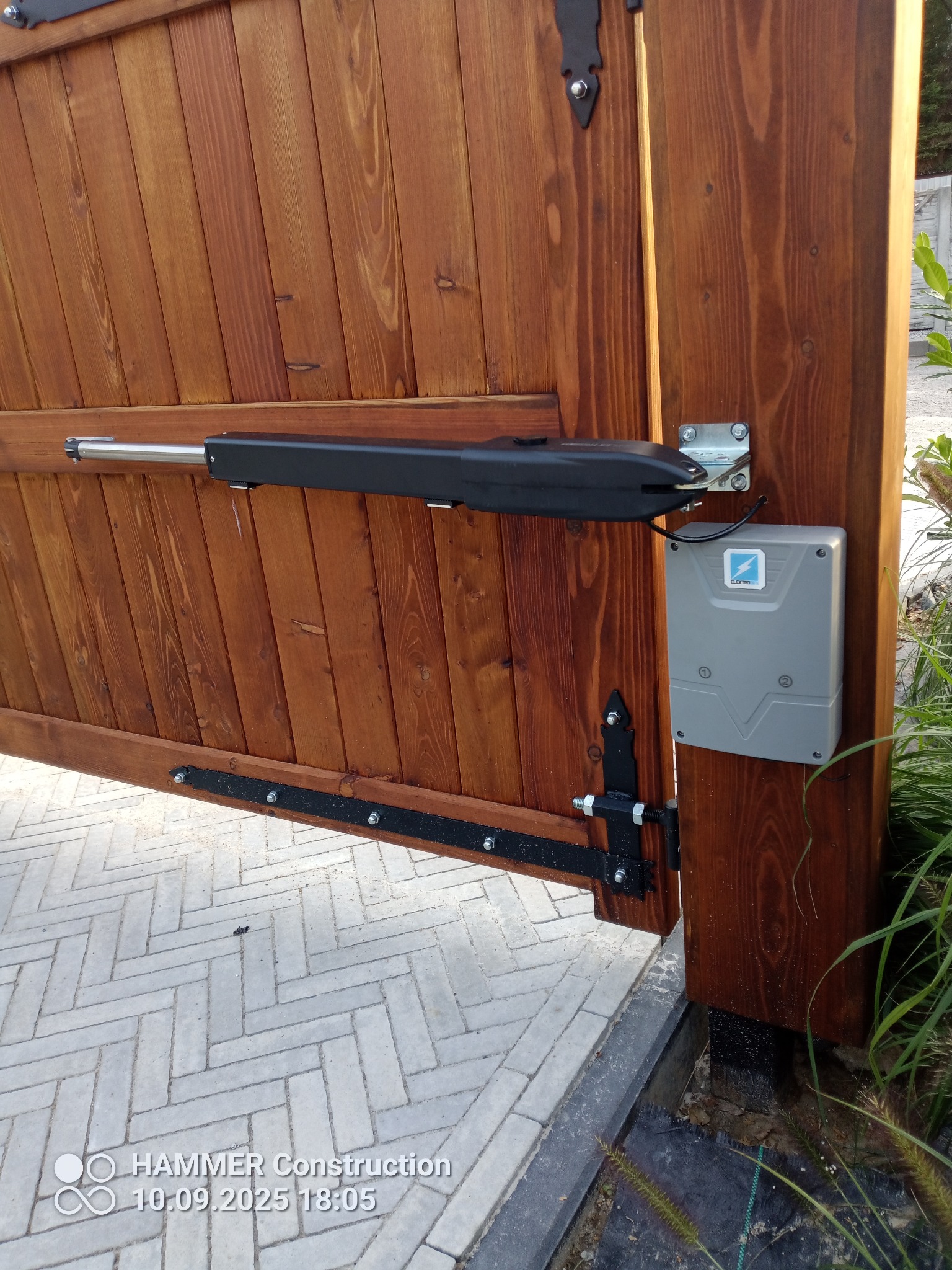 Drewniana brama z zamontowaną automatyką i sterownikiem. Widoczny siłownik i skrzynka sterująca z logo producenta. Brama otwarta, na pierwszym planie kostka brukowa.