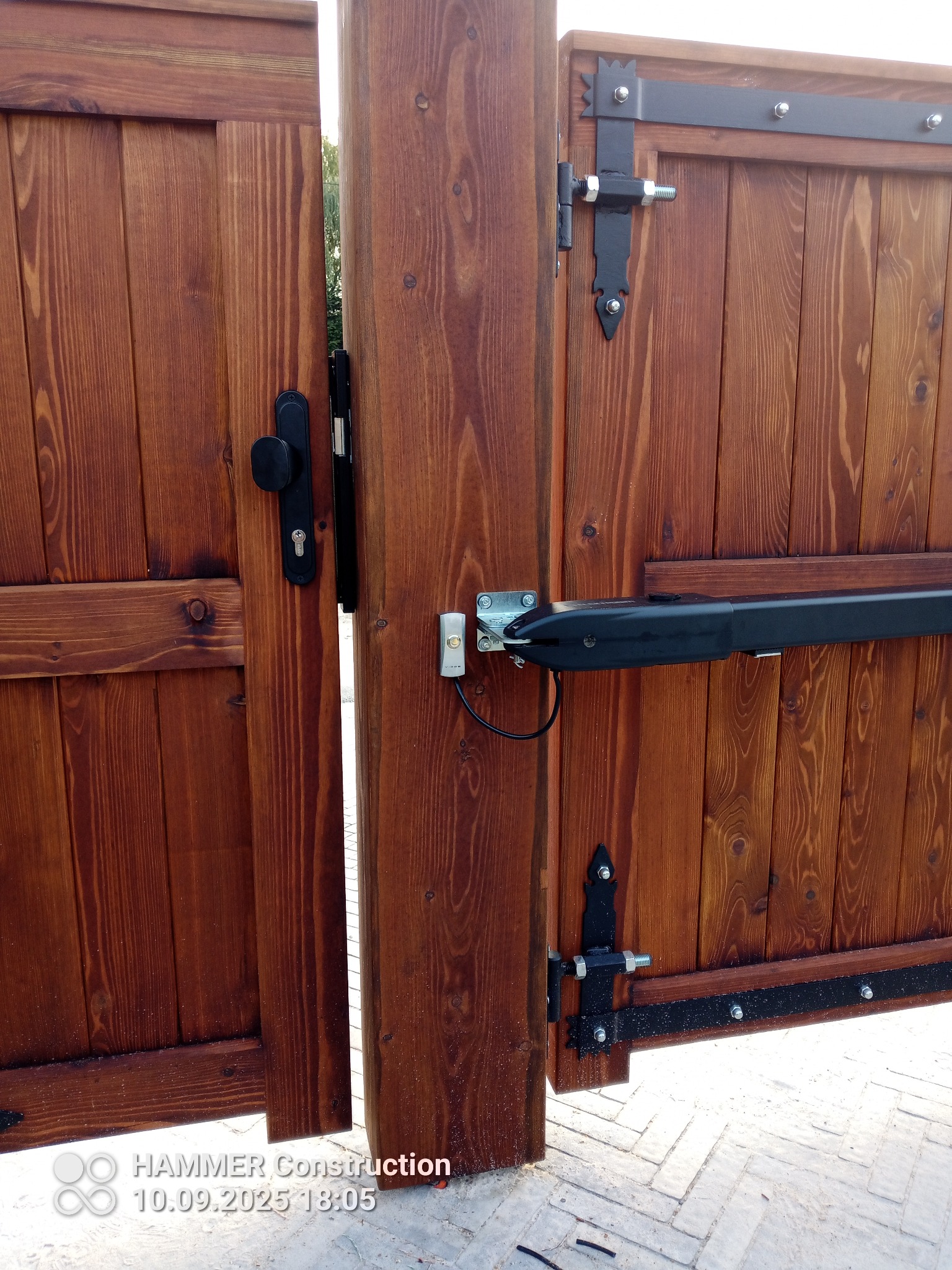 Drewniana brama dwuskrzydłowa z automatyką, widoczne zawiasy i zamek. Brama w kolorze brązowym, z wyraźnym rysunkiem słojów drewna. Elementy automatyki w kolorze czarnym i srebrnym.
