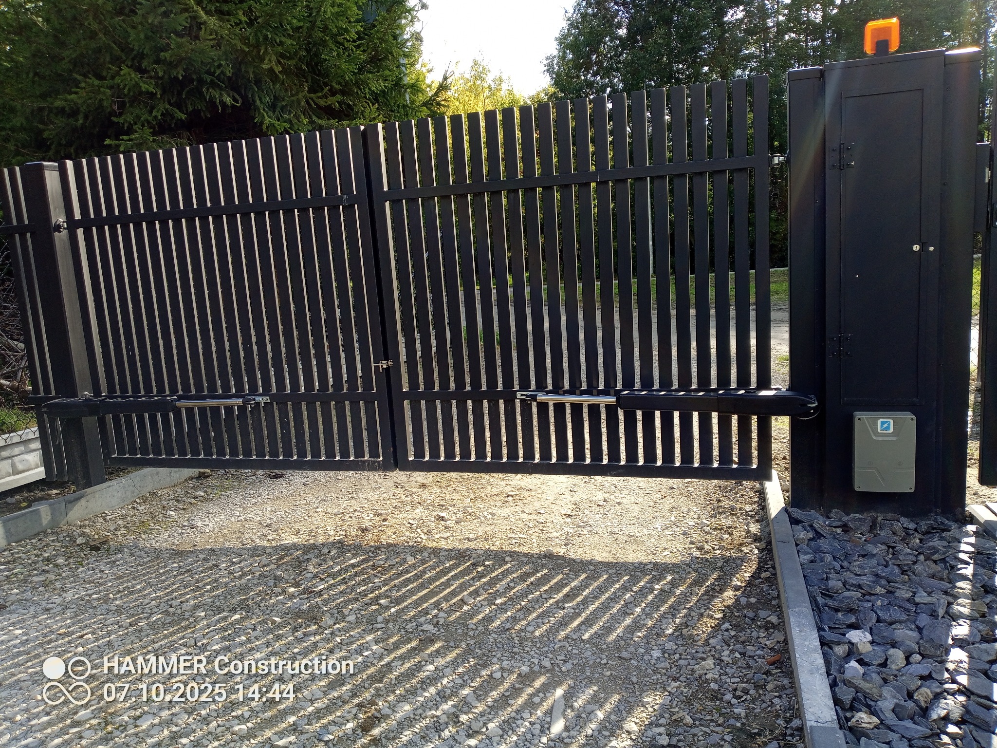 Nowoczesna, czarna brama automatyczna z pionowymi szczeblami, częściowo otwarta, z widocznym mechanizmem napędowym i lampą ostrzegawczą. Podłoże żwirowe.