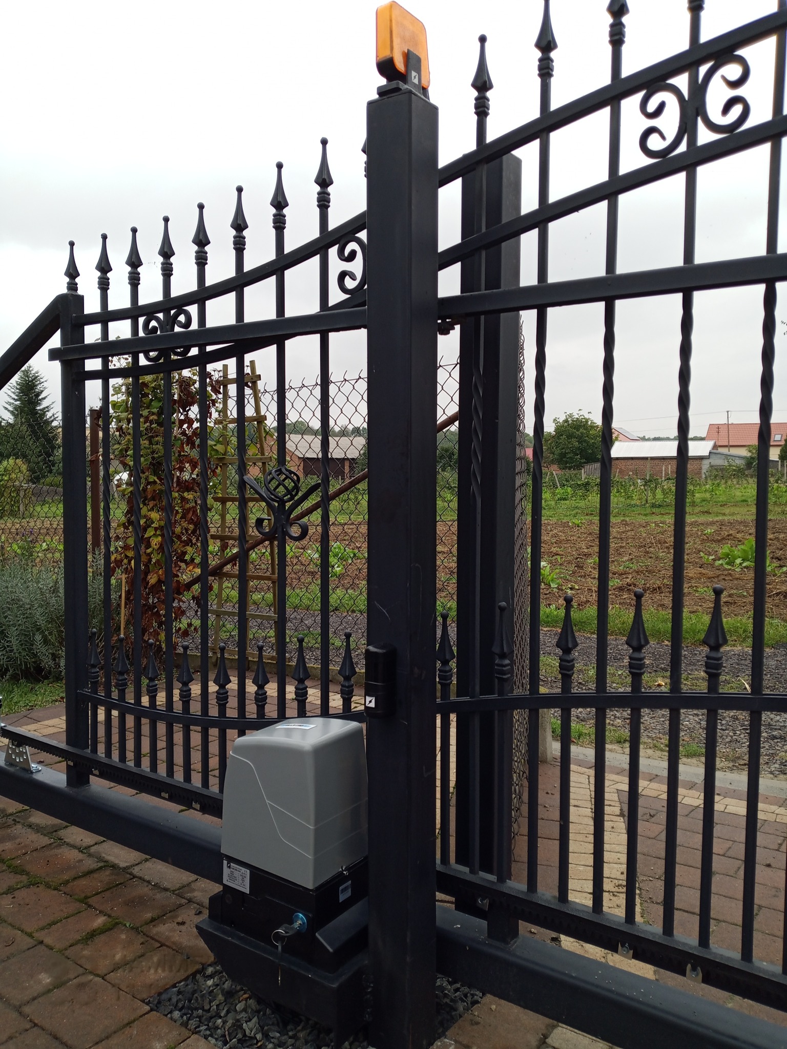 Kuta, czarna brama wjazdowa z automatyką, widoczny napęd i lampa ostrzegawcza. W tle ogród i zabudowania. Brama z ozdobnymi grotami i elementami dekoracyjnymi.