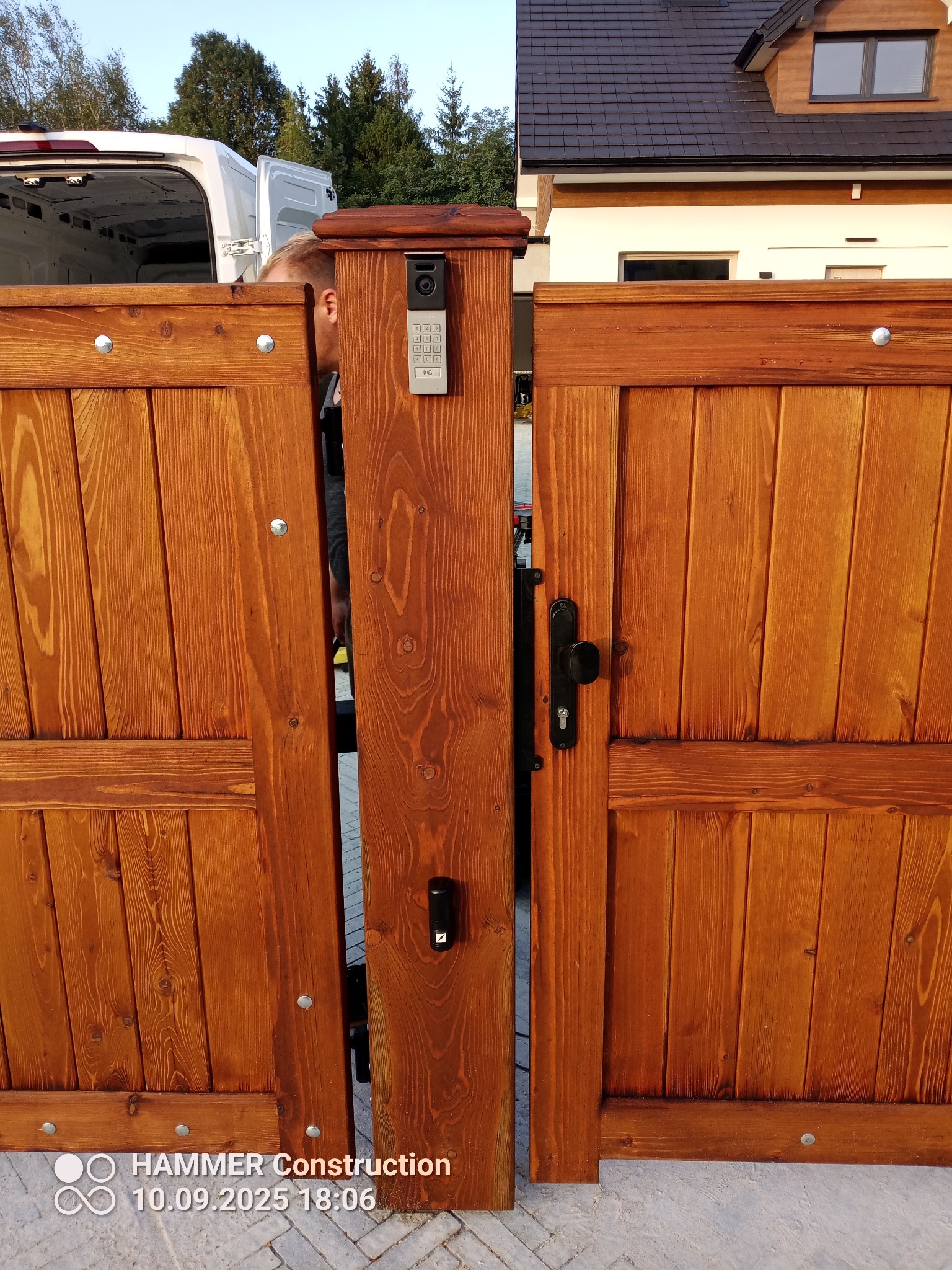 Drewniana brama wjazdowa z domofonem i panelem numerycznym, widoczny fragment budynku w tle. Brama w kolorze ciepłego brązu, z widoczną strukturą drewna.