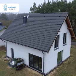 Marwel Home - domy prefabrykowane - Nowoczesny dom z białymi ścianami i grafitowym dachem z dachówki, widok z góry, otoczony zielenią. Widoczne duże okna i drzwi balkonowe.