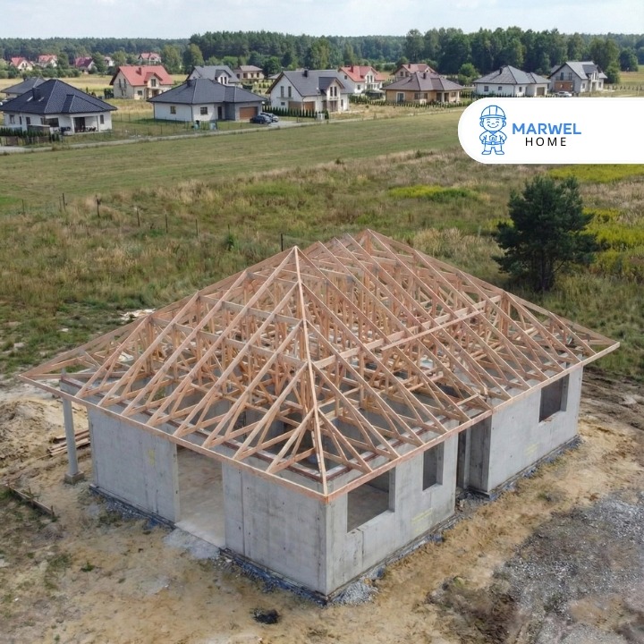 Surowy stan domu z betonu komórkowego z drewnianą konstrukcją dachu w trakcie budowy, na tle zieleni i zabudowy jednorodzinnej. Widok z lotu ptaka.