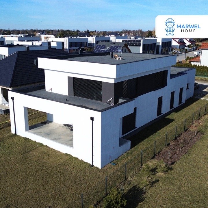 Nowoczesny, biały dom z płaskim dachem i dużymi oknami, otoczony zielonym trawnikiem i ogrodzeniem. Widok z góry, w tle inne domy i błękitne niebo.