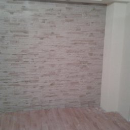 Ściana wykończona dekoracyjną, beżową okładziną imitującą kamień, z widoczną podłogą z paneli w odcieniach brązu. Fragment narożnika pokoju z kolumną.