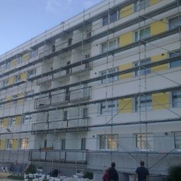 Wilk - Budynek wielorodzinny w trakcie prac elewacyjnych, otoczony rusztowaniami. Widoczne puszki z farbą i dwie osoby u dołu kadru. Częściowo pomalowana fasada na biało i żółto.