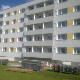 Wilk - Wielopiętrowy budynek z balkonami i żółtymi pasami elewacji. Na trawniku składowane metalowe elementy, prawdopodobnie rusztowania.