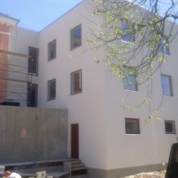 Wilk - Otynkowany budynek z oknami, częściowo w rusztowaniach, z widocznymi detalami wykończenia elewacji i elementami krajobrazu, jak gałęzie drzew z młodymi liśćmi.