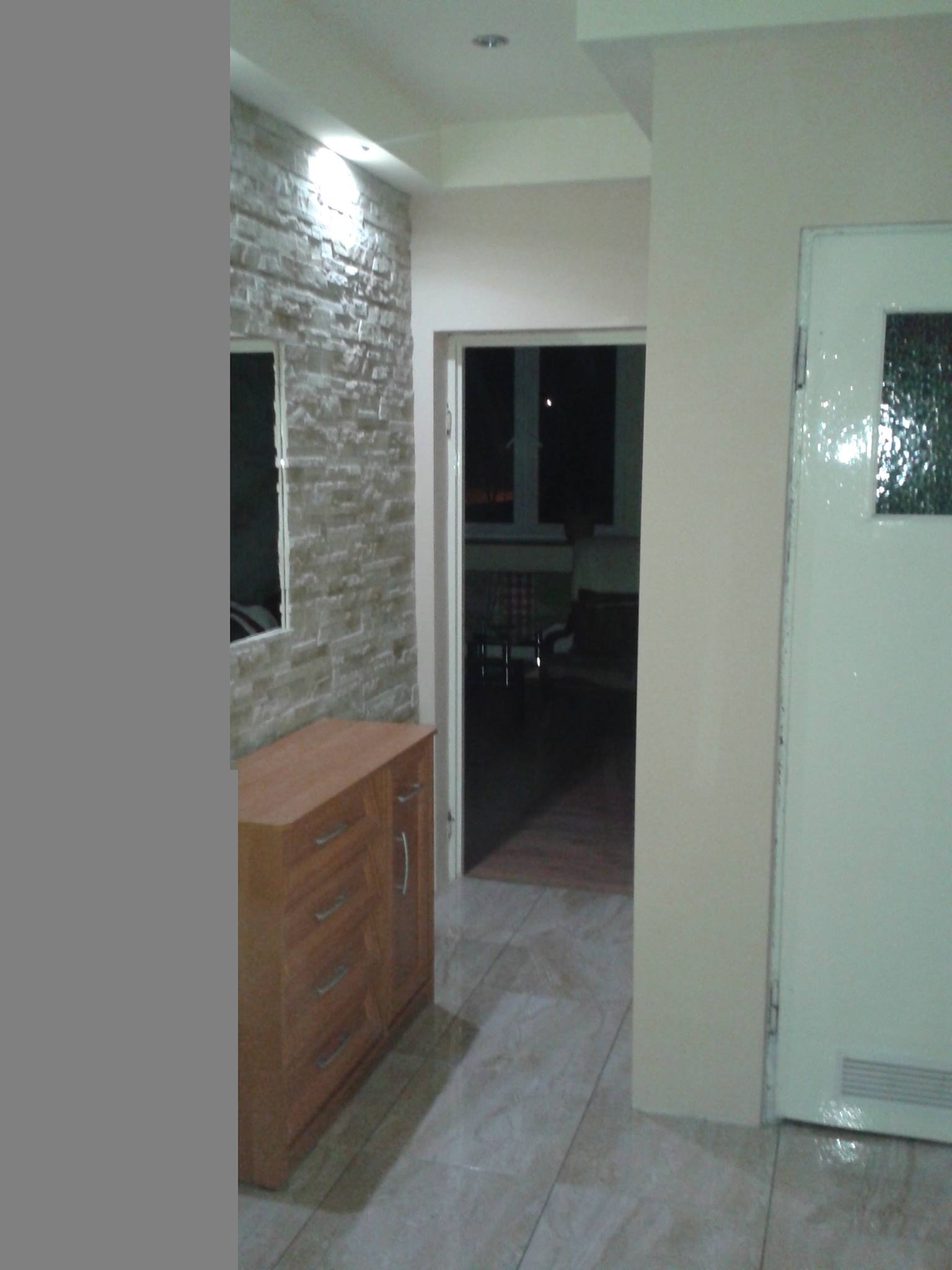 Korytarz po remoncie: ściana z dekoracyjnej cegły, drewniana komoda z lustrem, jasna podłoga z płytek, białe drzwi z szybą. Widok na pokój przez otwarte drzwi.