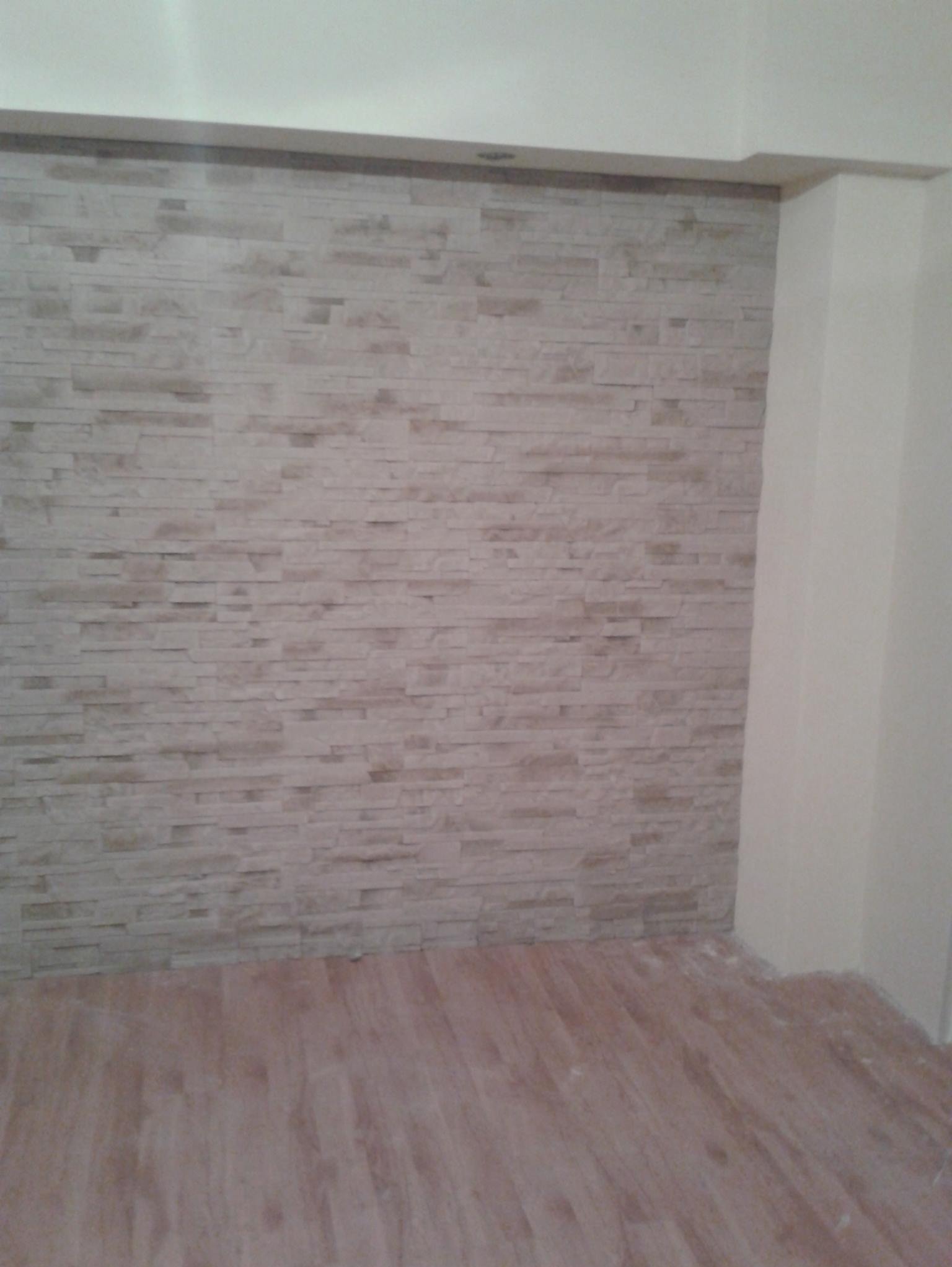 Ściana wykończona dekoracyjną, beżową okładziną imitującą kamień, z widoczną podłogą z paneli w odcieniach brązu. Fragment narożnika pokoju z kolumną.