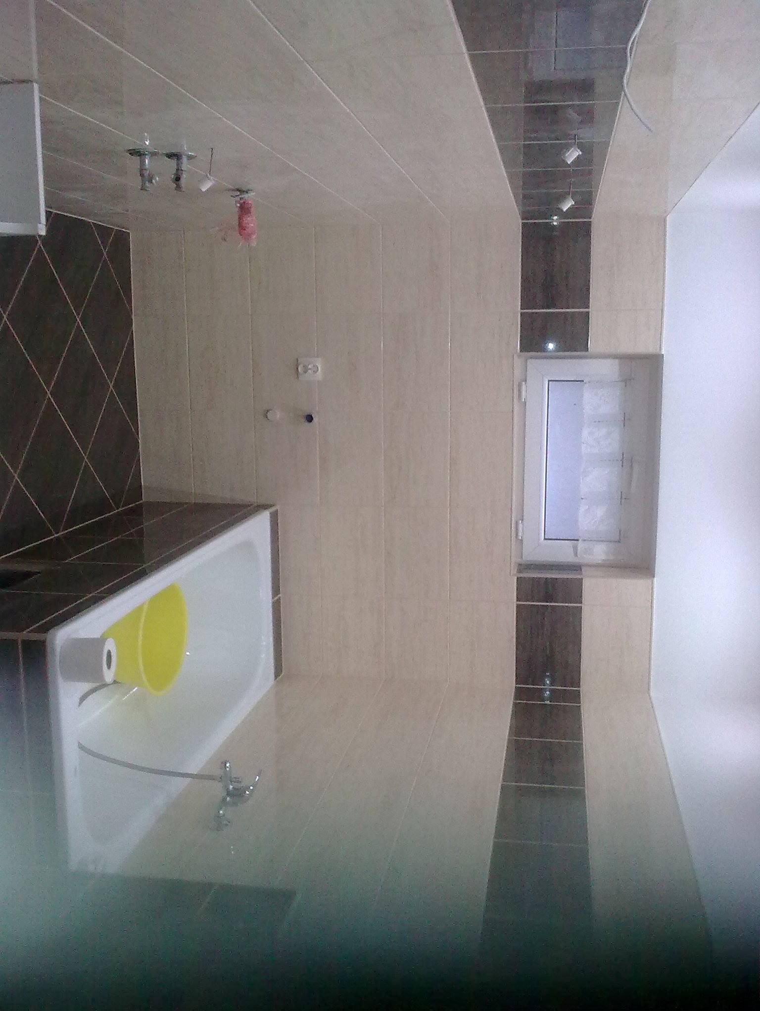 Wykończona łazienka z beżowymi i brązowymi płytkami, białą wanną z żółtą miską i oknem. Widoczne oświetlenie punktowe i armatura. Ujęcie z niskiego kąta.