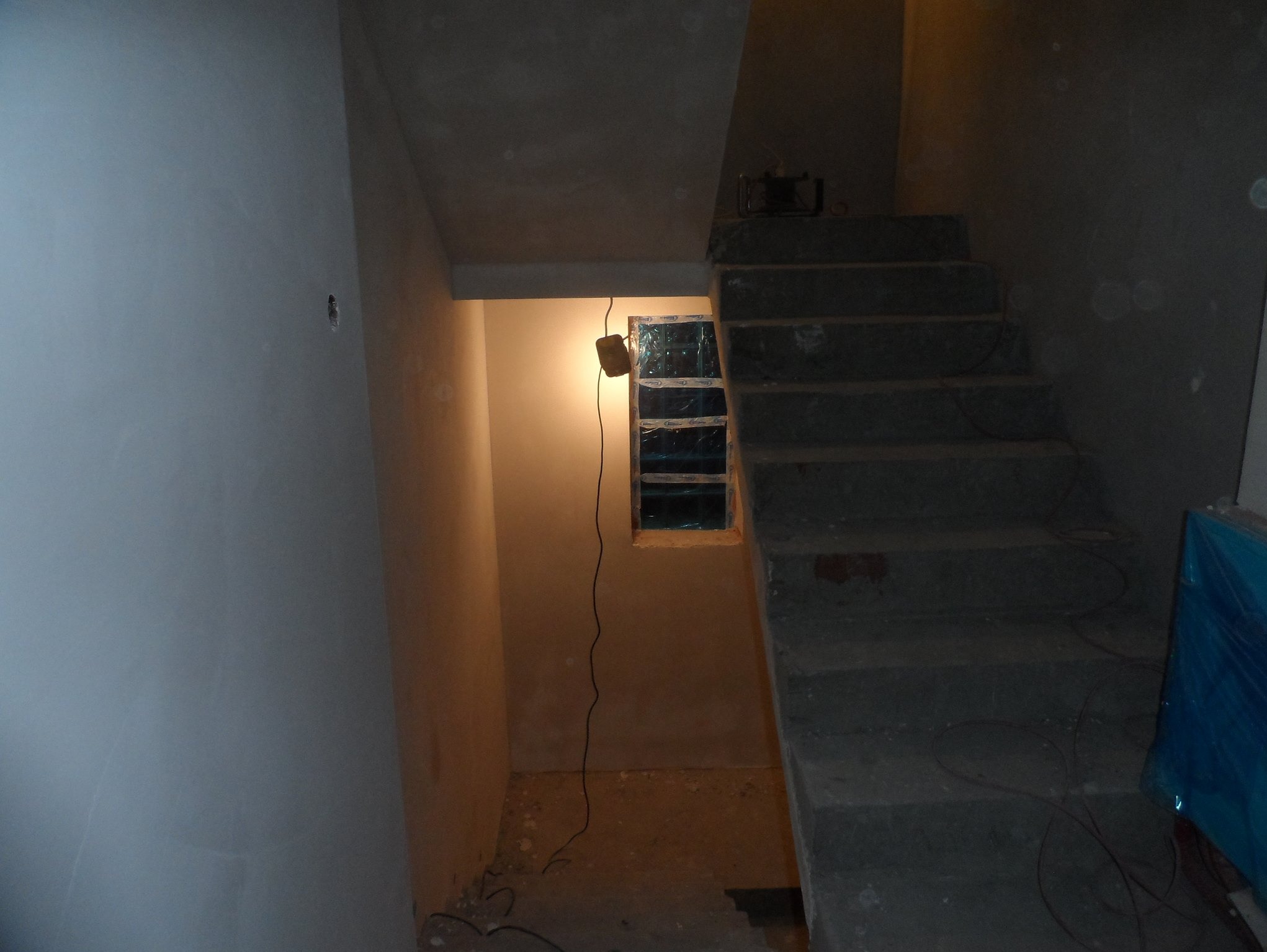 Surowe schody betonowe w trakcie budowy, zabezpieczone okno, kabel zwisający ze ściany, surowy tynk. Wnętrze w stanie deweloperskim, oświetlone lampą roboczą.