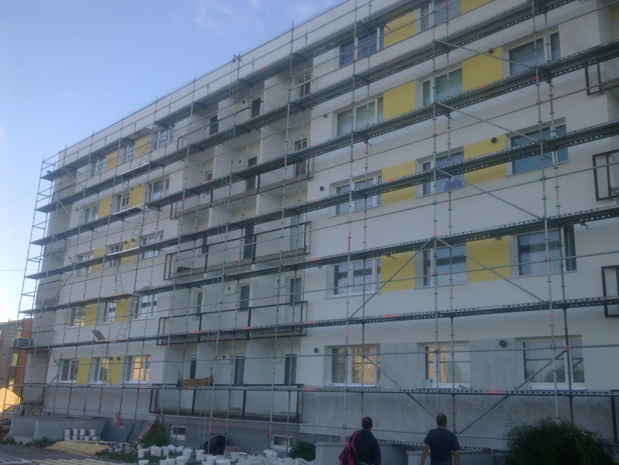Budynek wielorodzinny w trakcie prac elewacyjnych, otoczony rusztowaniami. Widoczne puszki z farbą i dwie osoby u dołu kadru. Częściowo pomalowana fasada na biało i żółto.