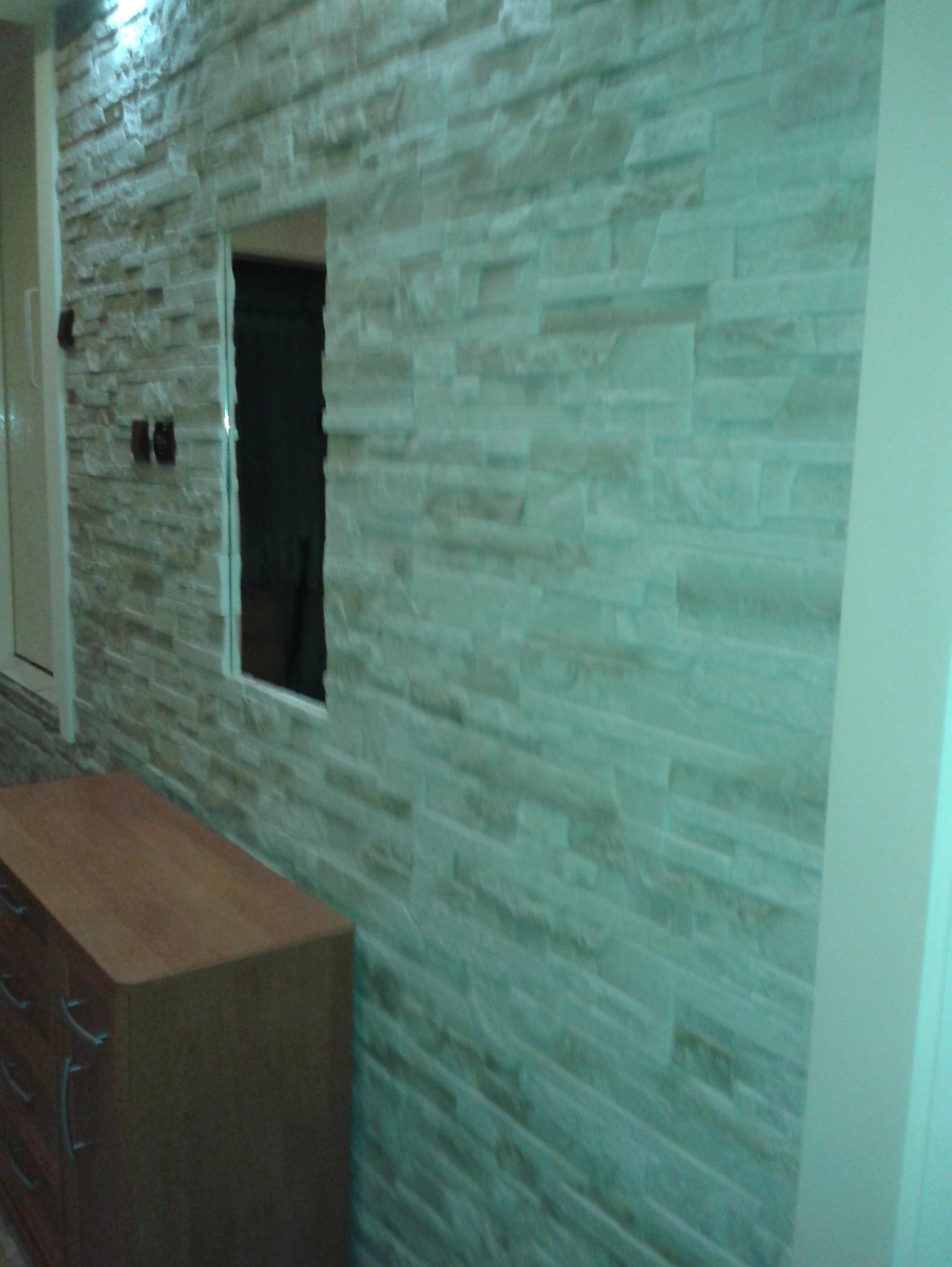 Ściana wykończona dekoracyjną okładziną imitującą kamień, z widocznym otworem i fragmentem komody. Neutralna kolorystyka, nierówna faktura.