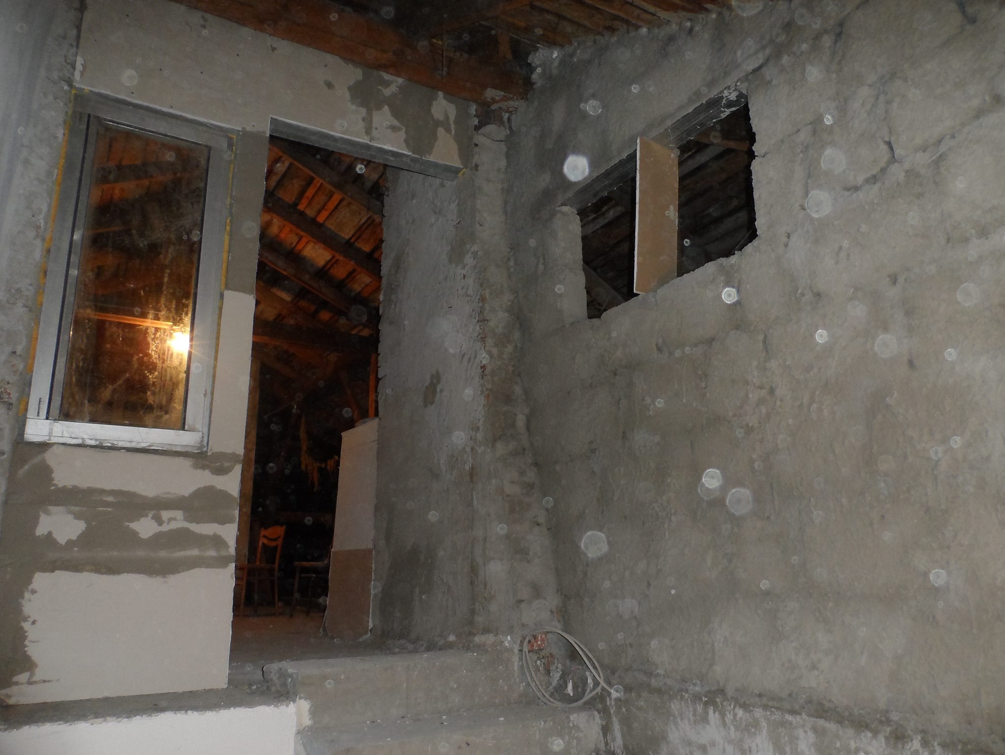 Surowy stan wnętrza budynku w trakcie prac wykończeniowych: widoczne okna w trakcie montażu, ściany w stanie deweloperskim, fragment więźby dachowej w tle.