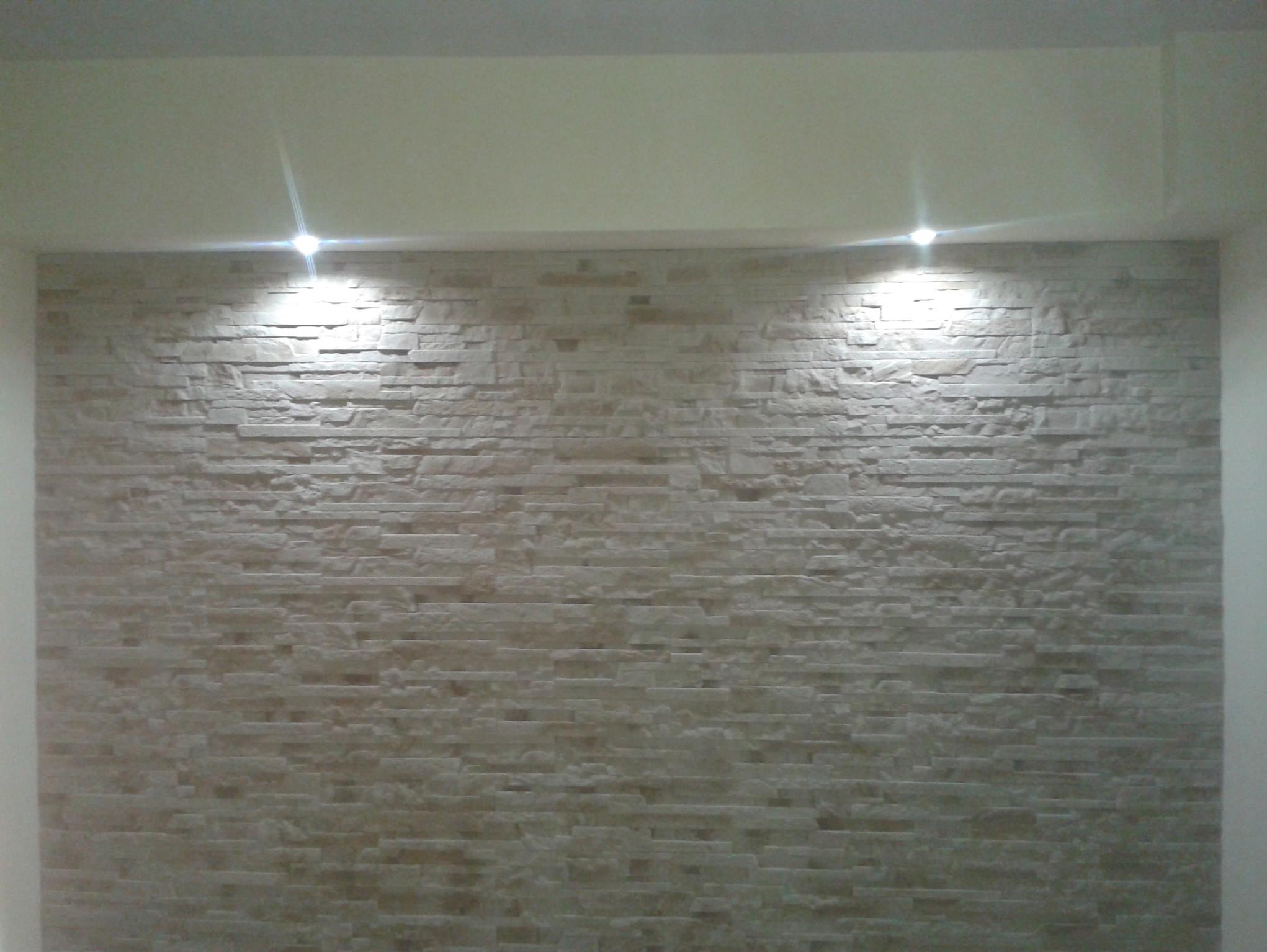 Ściana wykończona dekoracyjną, beżową okładziną imitującą kamień, oświetlona dwoma punktowymi lampami sufitowymi. Widok na całą ścianę, bez elementów wyposażenia.