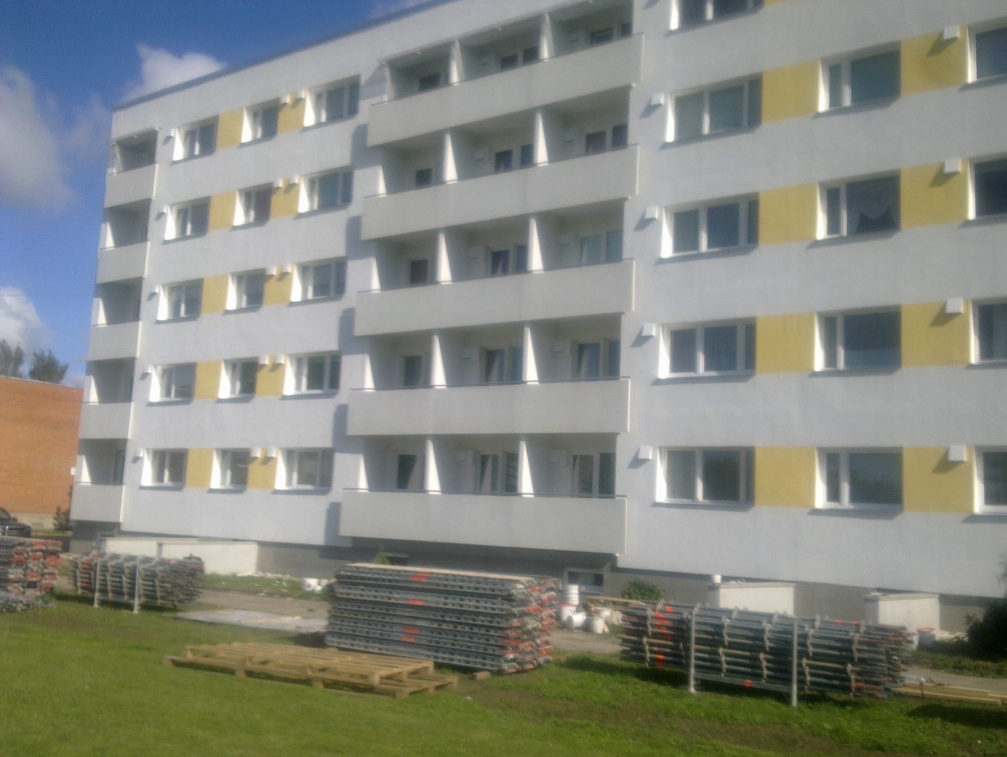 Wielopiętrowy budynek z balkonami i żółtymi pasami elewacji. Na trawniku składowane metalowe elementy, prawdopodobnie rusztowania.