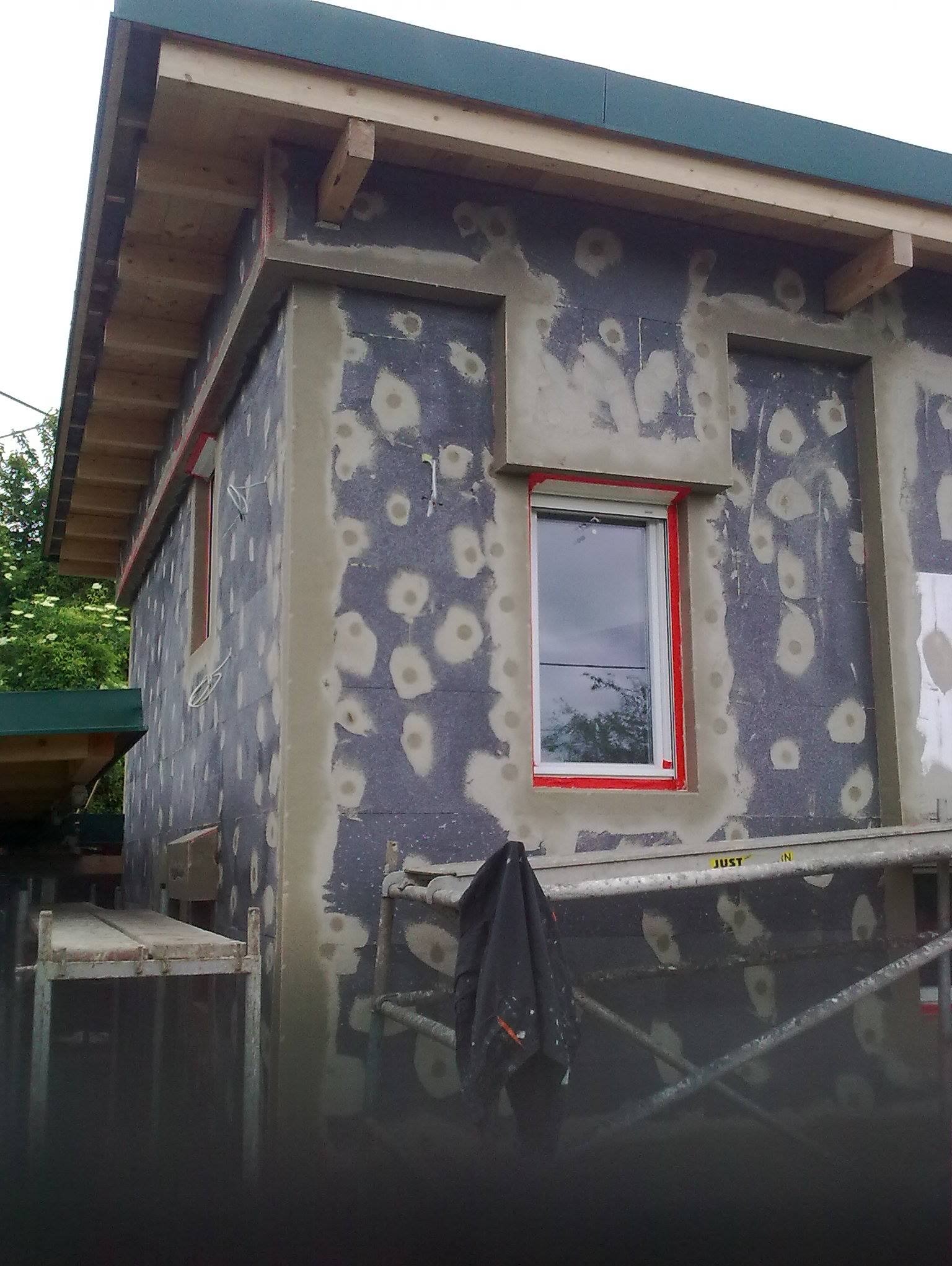 Budynek w trakcie ocieplania styropianem grafitowym, widoczne szpachlowane kołki mocujące. Okna z czerwoną taśmą zabezpieczającą. Rusztowanie przy ścianie.