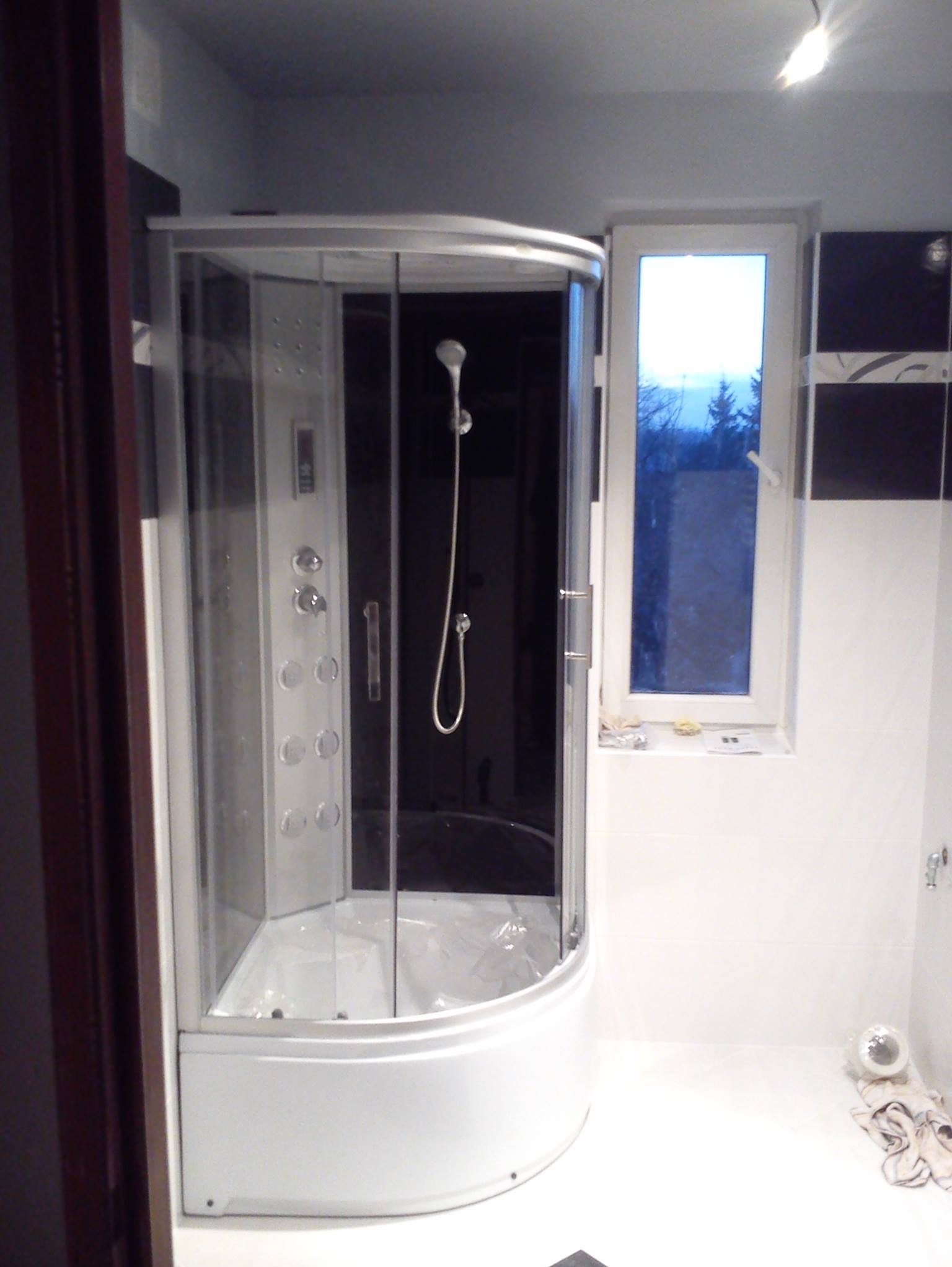 Nowoczesna kabina prysznicowa z hydromasażem w jasnej łazience z oknem. Czarne płytki akcentują ścianę. Widoczne detale wykończenia i armatura.