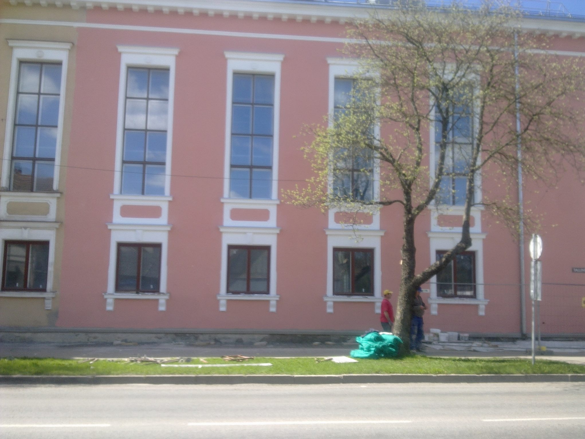 Różowa fasada budynku z białymi obramowaniami okien, częściowo zasłonięta przez drzewo. Dwóch robotników stoi obok worków z materiałami budowlanymi.