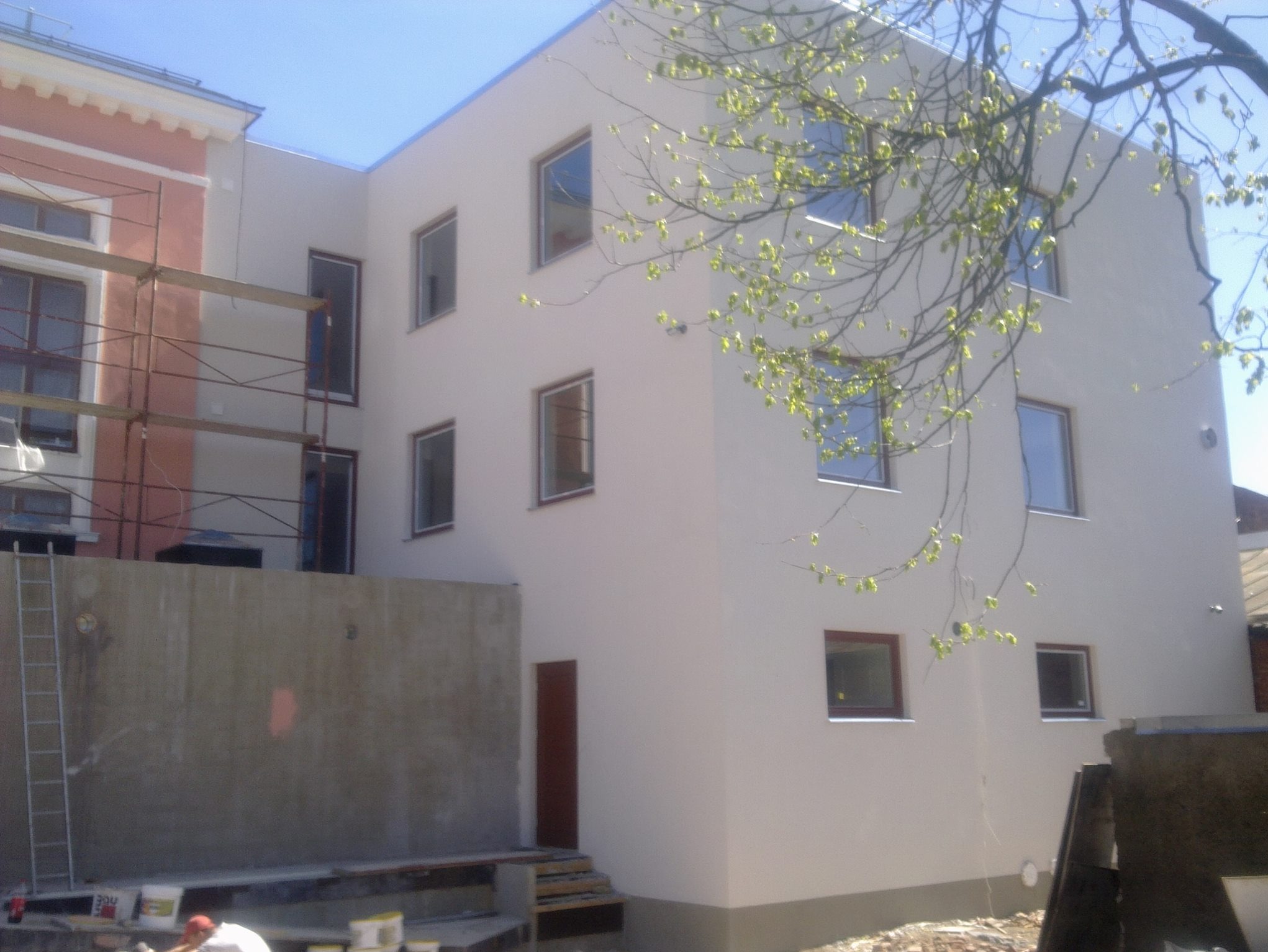 Otynkowany budynek z oknami, częściowo w rusztowaniach, z widocznymi detalami wykończenia elewacji i elementami krajobrazu, jak gałęzie drzew z młodymi liśćmi.