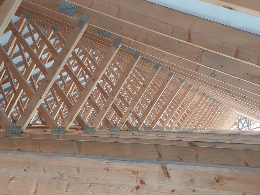 Drewniana konstrukcja dachu z widocznymi wiązarami kratowymi i metalowymi łącznikami, ukazana z perspektywy wnętrza budynku w trakcie budowy.