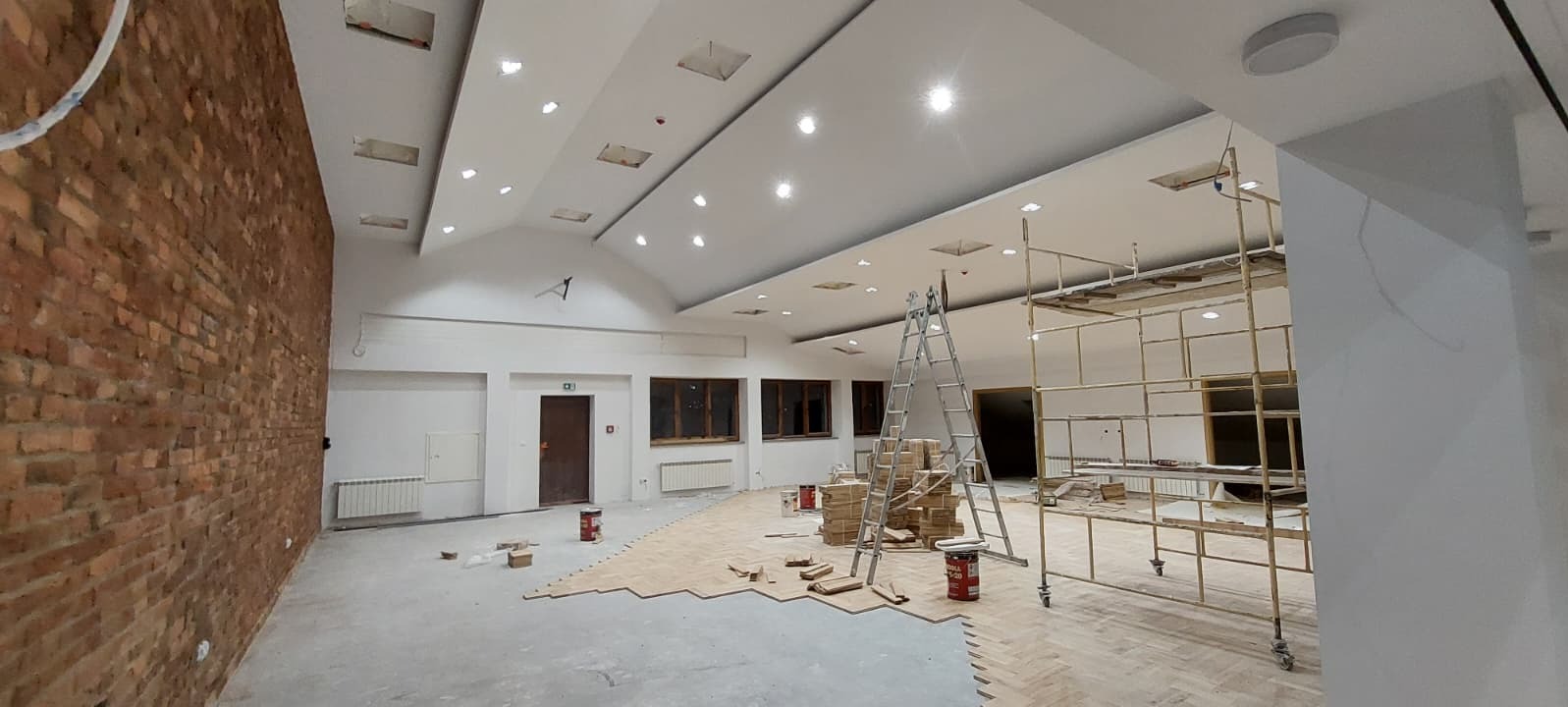 Wnętrze w trakcie wykończenia: częściowo ułożony parkiet w jodełkę, ceglana ściana, rusztowania, puszki z farbą. Sufit z oświetleniem punktowym. Widok na okna i drzwi.