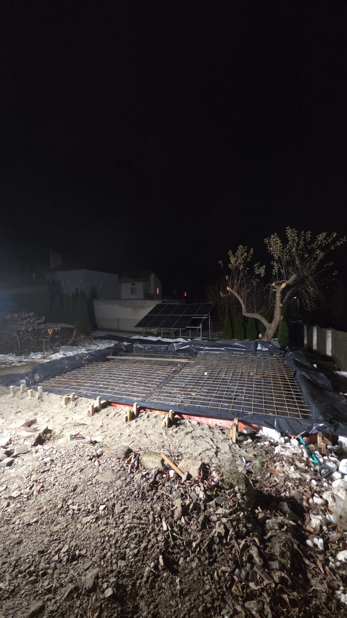 Nocne ujęcie z ziemi: przygotowany fundament z siatką zbrojeniową, deskowaniem i folią, gotowy do zalania betonem. W tle dom i instalacja solarna.