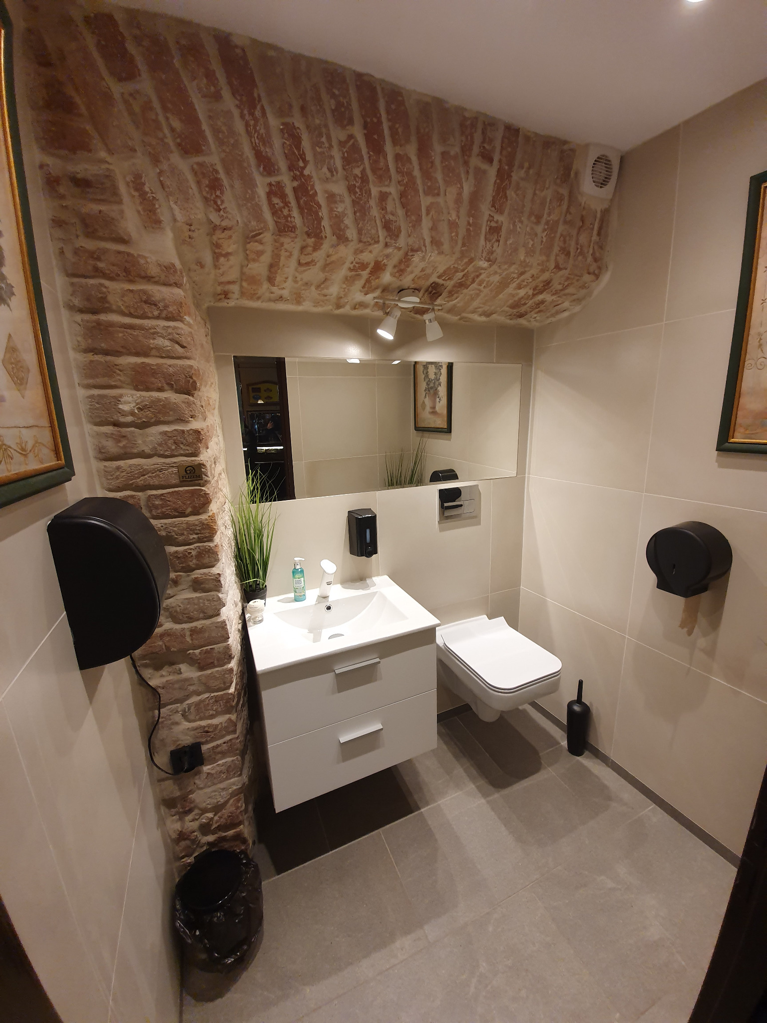 Nowoczesna toaleta z elementami cegły: umywalka z szafką, wisząca muszla, szara podłoga. Nad umywalką lustro, a nad nim ceglane sklepienie. Dystrybutor mydła i ręczników.